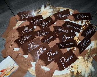 Op de afbeelding: Bruine naamkaartjes met witte kalligrafie namen, gerangschikt op een geweven basis met gedroogde bloemen en bruine linten. Namen zijn Delphine, Alex en Tatiana. Perfect voor bruiloften.
