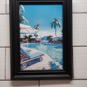 May include: A framed print showing a resort scene with a swimming pool, palm trees, and sun loungers. The artwork is in a black rectangular frame. The image depicts a sunny day with a blue sky and clear water.