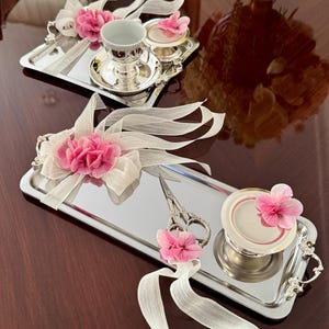 May include: Two silver trays with decorative elements. Each tray features a pink flower arrangement with white ribbon. One tray holds a teacup and saucer, the other holds a small container and scissors. The trays are on a dark brown surface.