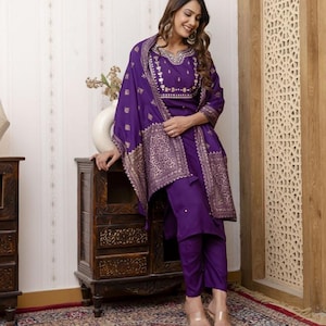 May include: A purple embroidered salwar kameez set. The set includes a tunic, pants, and a shawl. The tunic and shawl feature gold-colored embroidery. The outfit is worn with clear sandals. The ensemble is suitable for festive occasions.