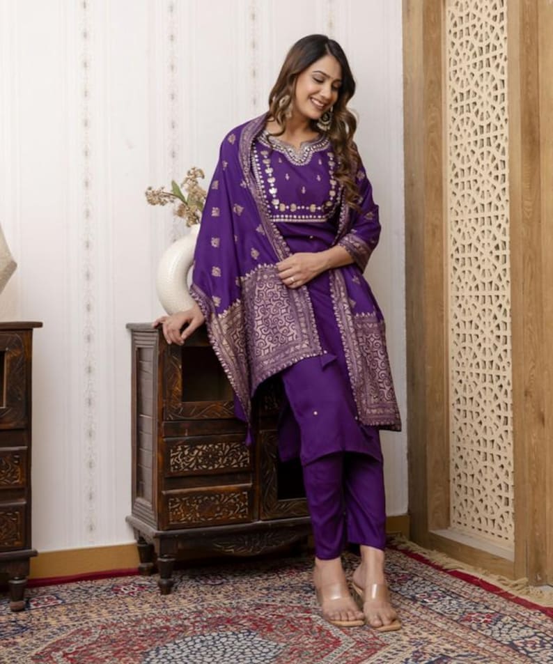 May include: A purple embroidered salwar kameez set. The set includes a tunic, pants, and a shawl. The tunic and shawl feature gold-colored embroidery. The outfit is worn with clear sandals. The ensemble is suitable for festive occasions.