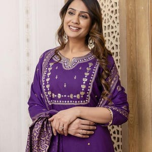 May include: A purple Indian suit with gold embroidery. The suit includes a long-sleeved tunic and matching scarf. The gold embroidery is on the neckline, sleeves, and scarf. The outfit is made of a soft fabric.