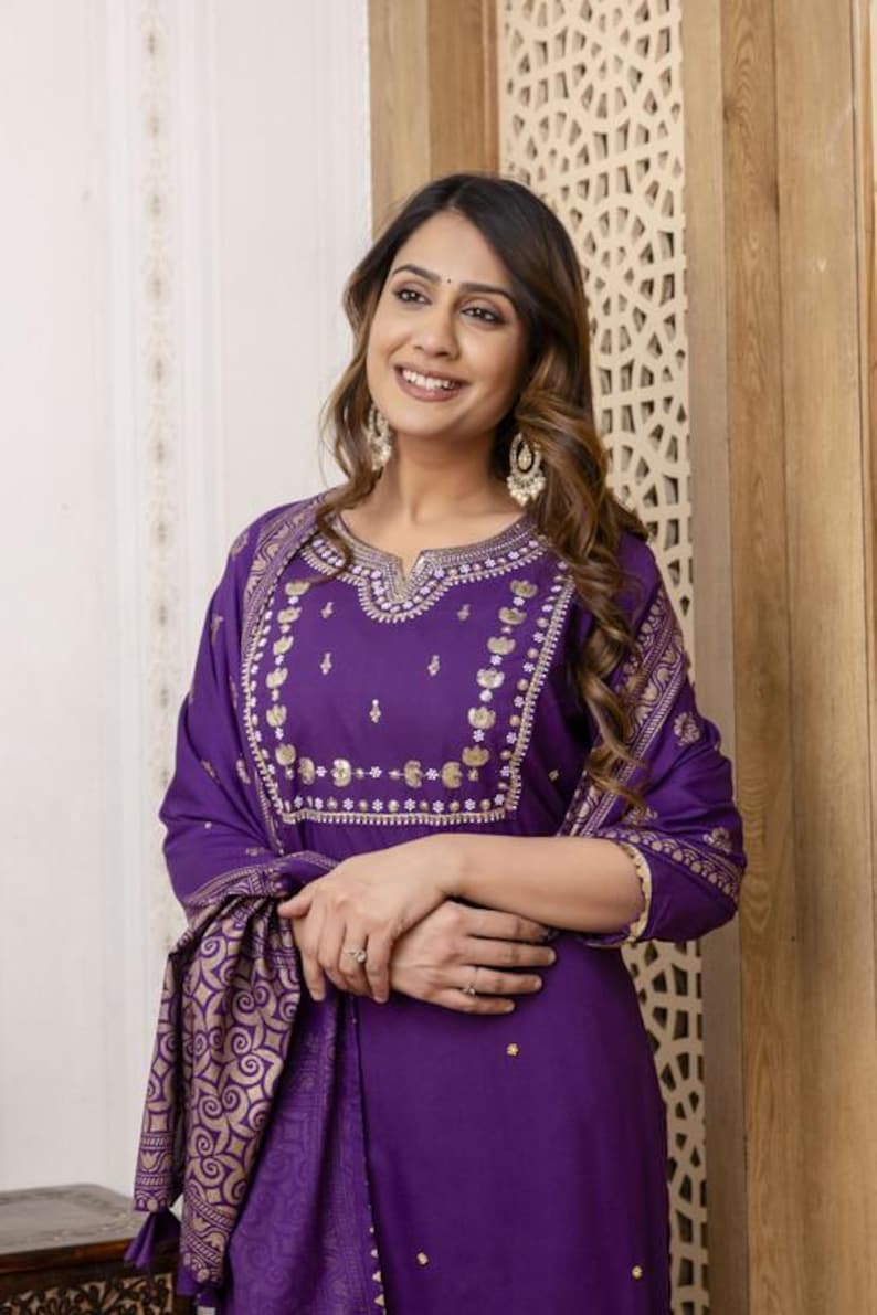 May include: A purple Indian suit with gold embroidery. The suit includes a long-sleeved tunic and matching scarf. The gold embroidery is on the neckline, sleeves, and scarf. The outfit is made of a soft fabric.