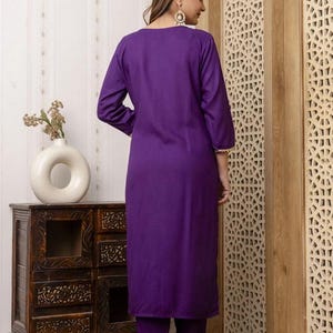 May include: A vibrant purple kurta suit. The long-sleeved tunic and matching pants are a solid color. The outfit is worn with clear heeled sandals. The background includes a wooden cabinet and a decorative wooden panel.