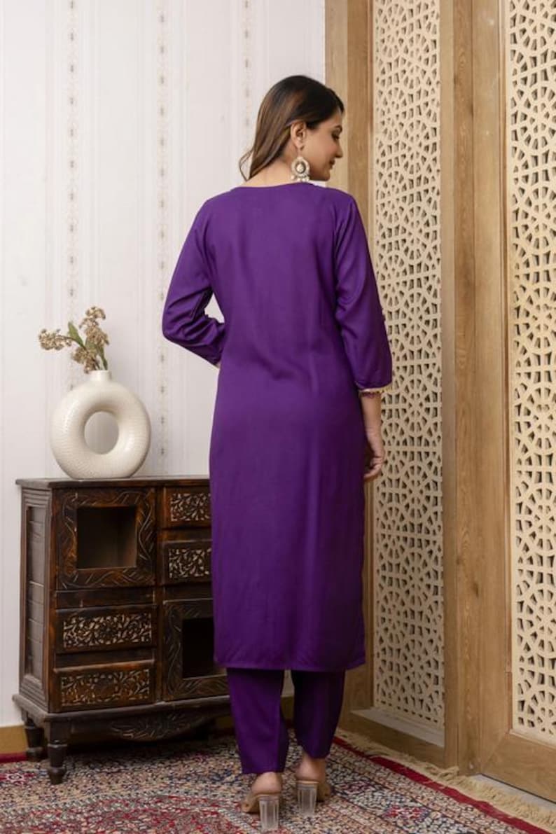 May include: A vibrant purple kurta suit. The long-sleeved tunic and matching pants are a solid color. The outfit is worn with clear heeled sandals. The background includes a wooden cabinet and a decorative wooden panel.