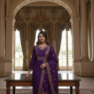 May include: A woman in a purple salwar kameez with gold embroidery and a matching dupatta stands near a table. The outfit features intricate gold patterns. The background includes a chandelier, ornate architecture, and a patterned rug.