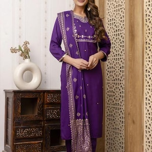 May include: A woman wearing a purple salwar kameez suit with gold embroidery. The long-sleeved tunic and matching pants are paired with a patterned scarf. The outfit is a traditional Indian style.