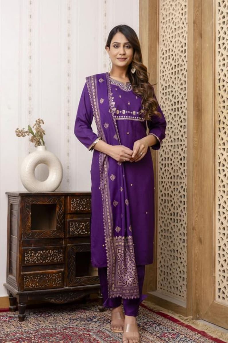 May include: A woman wearing a purple salwar kameez suit with gold embroidery. The long-sleeved tunic and matching pants are paired with a patterned scarf. The outfit is a traditional Indian style.