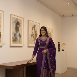 May include: A woman in a purple embroidered salwar kameez, a traditional South Asian outfit, stands in an art gallery. The outfit includes a long-sleeved tunic, matching pants, and a patterned scarf. The ensemble is adorned with gold embroidery.