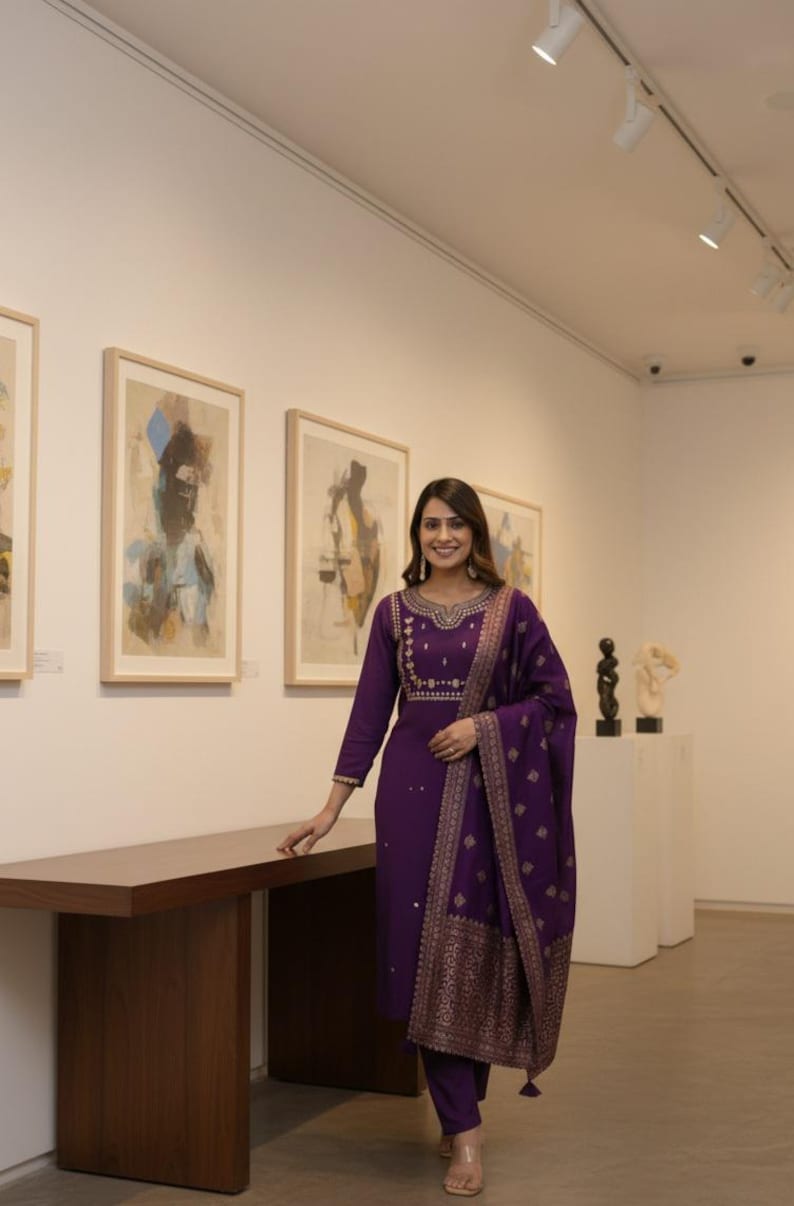 May include: A woman in a purple embroidered salwar kameez, a traditional South Asian outfit, stands in an art gallery. The outfit includes a long-sleeved tunic, matching pants, and a patterned scarf. The ensemble is adorned with gold embroidery.