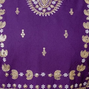 May include: Close-up of a purple garment with intricate gold embroidery and white accents. The design features floral and geometric patterns along the neckline and edges, creating a decorative and elegant look.
