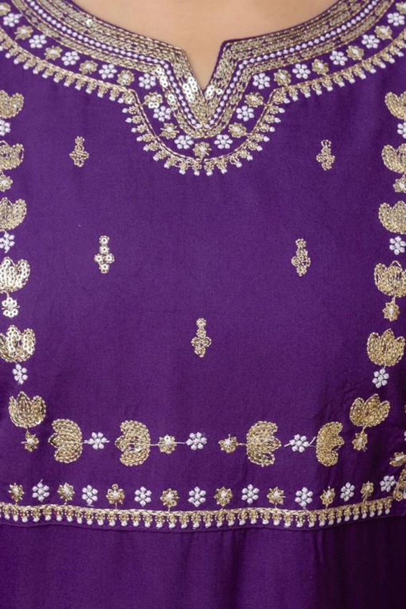 May include: Close-up of a purple garment with intricate gold embroidery and white accents. The design features floral and geometric patterns along the neckline and edges, creating a decorative and elegant look.