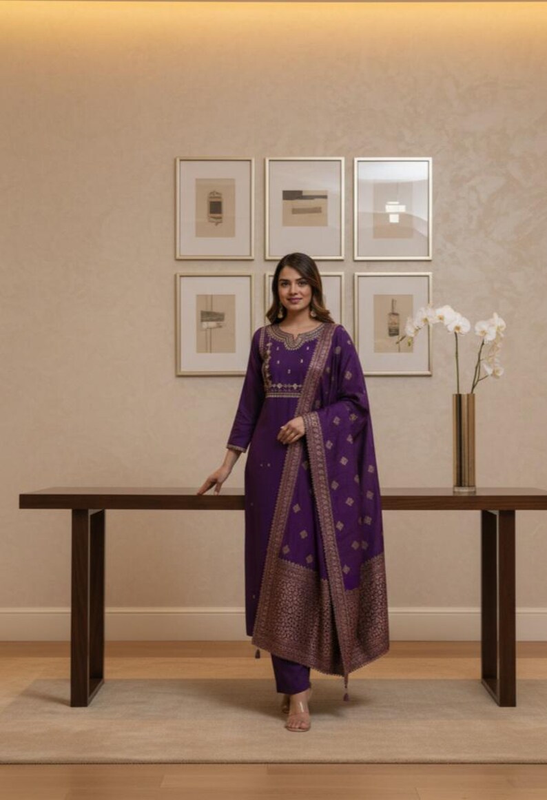May include: A woman wearing a purple salwar kameez with gold embroidery and a matching dupatta. The outfit features intricate patterns and is paired with matching pants. The background includes framed artwork and a wooden table, creating an elegant setting.