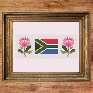 May include: Cross-stitch artwork featuring the South African flag and two pink protea flowers. The design is framed in a gold-colored frame, set against a wooden background. The words "Instant Download PDF" are in the bottom right corner.