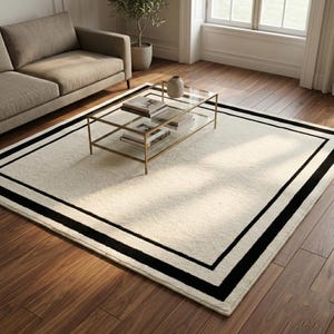 May include: A rectangular area rug featuring a cream-colored center with a double black border frame. The rug is positioned on a wooden floor beneath a glass coffee table with a gold metal frame and a beige sofa in the background.