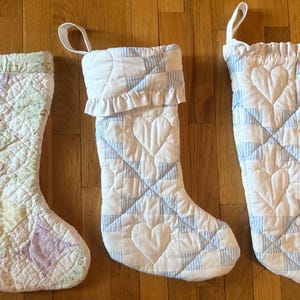 May include: Three quilted Christmas stockings with hanging loops. The left stocking features a pastel patchwork pattern. The middle and right stockings are white with blue striped accents and quilted heart motifs, with the middle one featuring a ruffled cuff.