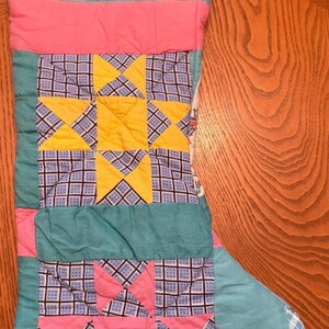 May include: A quilted Christmas stocking featuring a patchwork design with teal, pink, and yellow fabric panels. The center displays a yellow star pattern against a dark blue and white grid background, while the lower section has pink triangles.