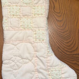 May include: A quilted Christmas stocking featuring a patchwork design of solid pale pink fabric squares and light green squares with a small floral pattern. The stocking includes a stitched circular floral motif and a white fabric hanging loop.