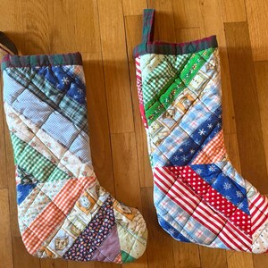 May include: Two quilted Christmas stockings constructed from a patchwork of various fabric scraps. The stockings feature diagonal patterns in red, blue, green, white, and orange, including gingham, polka dot, floral, and snowflake prints.