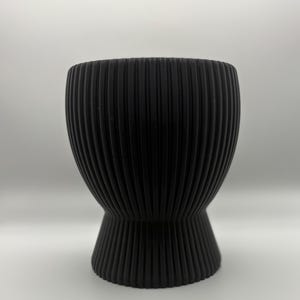 May include: A black, ribbed, decorative vase with a wide top and a flared base. The vase has a cylindrical shape with vertical grooves running from top to bottom. The vase is approximately 8 inches tall.