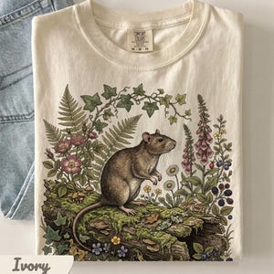 May include: An ivory short-sleeve t-shirt featuring a graphic print of a brown rat standing on a mossy log surrounded by ferns, ivy, pink flowers, foxgloves, daisies, and blackberries. The shirt has a Comfort Colors brand label at the neck.