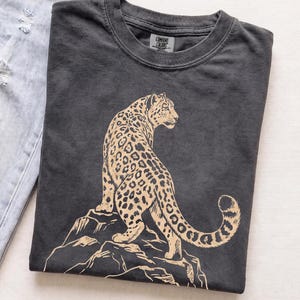 Folded charcoal Comfort Colors T-shirt featuring a retro golden snow leopard standing on rocky mountains, photographed on a light neutral background.