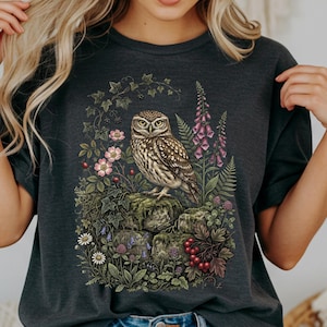 May include: A dark charcoal gray t-shirt featuring a detailed graphic print of a brown owl perched on mossy stones. The owl is surrounded by a botanical arrangement of pink flowers, purple foxgloves, green ferns, ivy, and clusters of red berries.