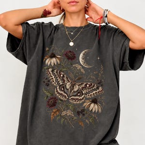 May include: A charcoal gray oversized t-shirt featuring a detailed graphic print of a large moth surrounded by wildflowers, berries, and a crescent moon. The design incorporates earthy tones of brown, cream, and muted green on the dark fabric.