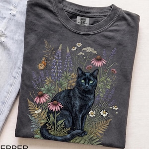 May include: A charcoal gray t-shirt featuring a graphic of a black cat sitting among colorful wildflowers, including purple lupines, pink coneflowers, and white daisies, with a monarch butterfly nearby. The shirt has a crew neck and a Comfort Colors label.