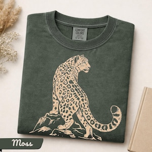 May include: A dark moss green crew neck t-shirt featuring a light beige graphic print of a leopard standing on a rock. The shirt has a ribbed collar and a Comfort Colors brand label printed on the inside of the neck area, indicating a size small.