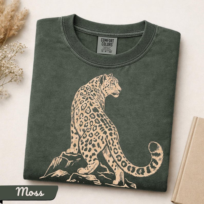 May include: A dark moss green crew neck t-shirt featuring a light beige graphic print of a leopard standing on a rock. The shirt has a ribbed collar and a Comfort Colors brand label printed on the inside of the neck area, indicating a size small.
