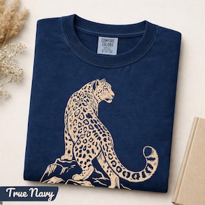 May include: A navy blue crew neck t-shirt featuring a light beige graphic of a leopard sitting on a rock. The interior neck label reads COMFORT COLORS and indicates size S. The shirt is folded neatly, displaying the animal illustration prominently.