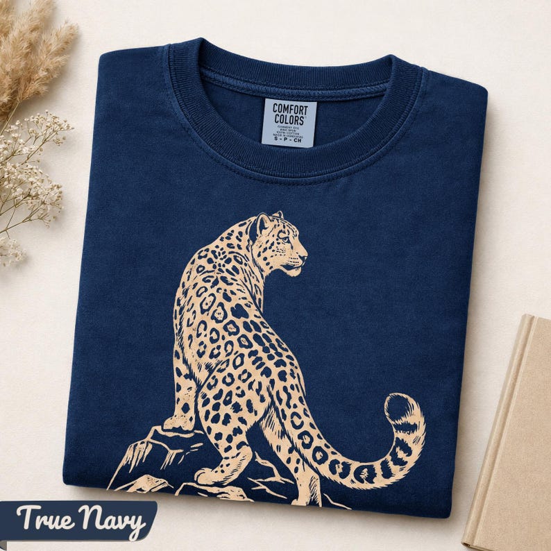 May include: A navy blue crew neck t-shirt featuring a light beige graphic of a leopard sitting on a rock. The interior neck label reads COMFORT COLORS and indicates size S. The shirt is folded neatly, displaying the animal illustration prominently.