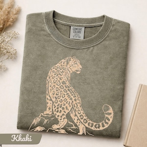 May include: A khaki green crew neck t-shirt featuring a light tan graphic print of a leopard sitting on a rock. The interior neck label displays the text COMFORT COLORS and size S-P-CH. The fabric has a washed, vintage-style texture.