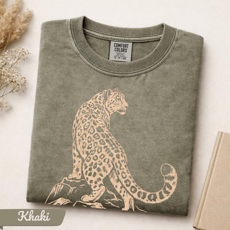 May include: A khaki green crew neck t-shirt featuring a light tan graphic print of a leopard sitting on a rock. The interior neck label displays the text COMFORT COLORS and size S-P-CH. The fabric has a washed, vintage-style texture.