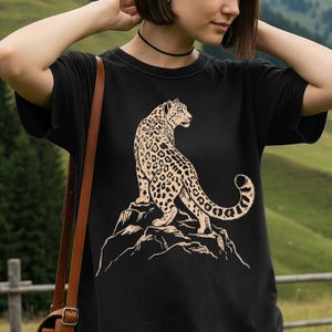 May include: A black short-sleeved t-shirt featuring a graphic print of a tan leopard standing on a rocky outcrop. The leopard is depicted in profile, looking toward the right, with its long tail curled upward. The design uses simple line work.