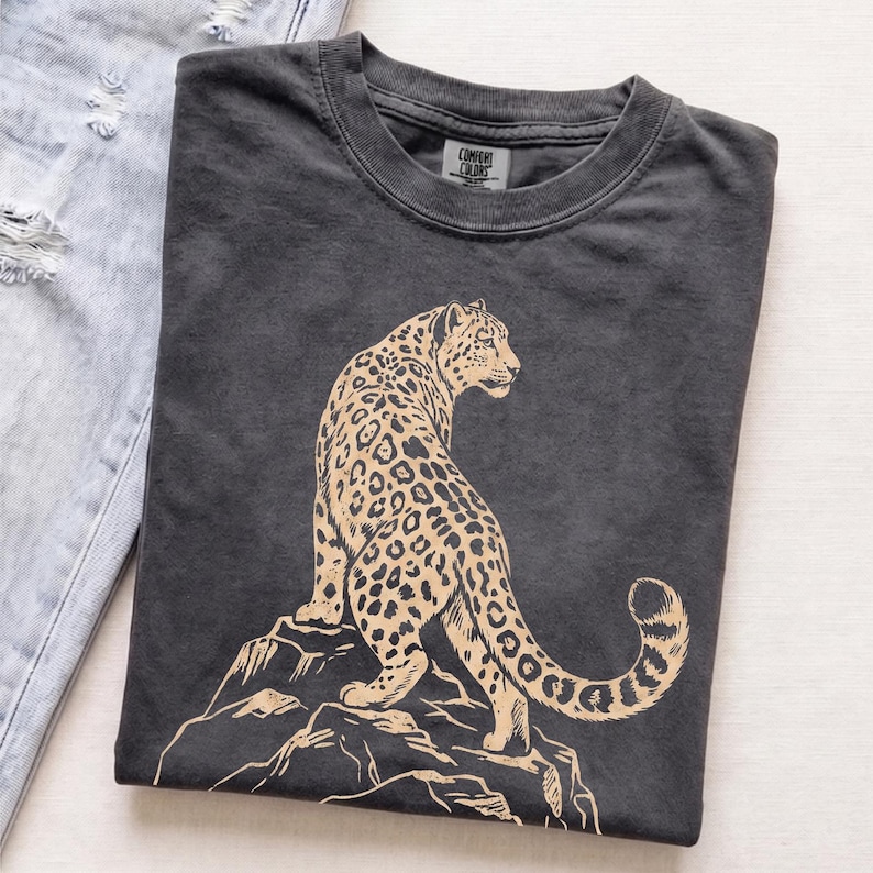 Folded charcoal Comfort Colors T-shirt featuring a retro golden snow leopard standing on rocky mountains, photographed on a light neutral background.