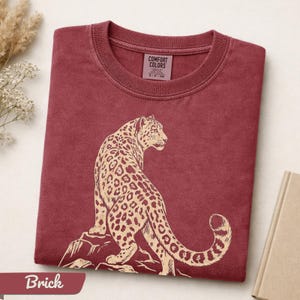 May include: A brick red crew neck t-shirt featuring a cream-colored graphic of a leopard sitting on a rock. The inner neck label displays the text Comfort Colors, 100% cotton, made in Honduras, and size S-P-CH. The fabric has a washed appearance.