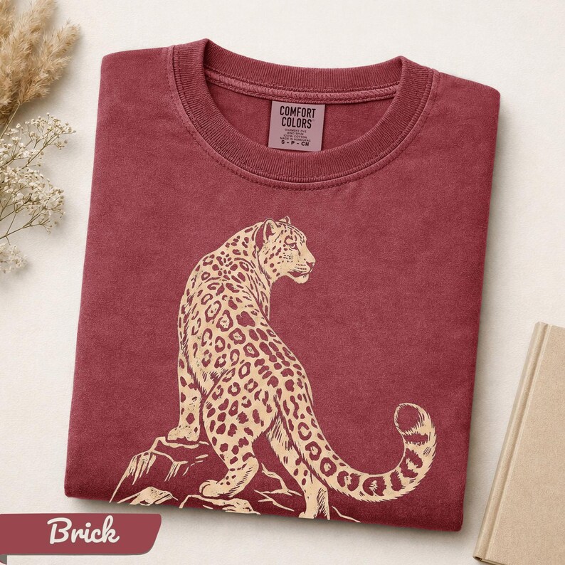May include: A brick red crew neck t-shirt featuring a cream-colored graphic of a leopard sitting on a rock. The inner neck label displays the text Comfort Colors, 100% cotton, made in Honduras, and size S-P-CH. The fabric has a washed appearance.
