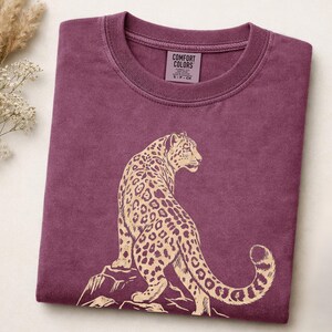 May include: A plum-colored crew neck t-shirt featuring a beige graphic print of a leopard sitting on a rock. The interior neck label displays the text COMFORT COLORS, S-P-CH, and additional garment information. The fabric has a washed texture.