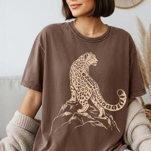 May include: A brown short-sleeved t-shirt featuring a graphic print of a leopard standing on a rocky outcrop. The leopard is depicted in a light tan color with dark brown spots, looking toward the right side of the shirt. The t-shirt has a crew neckline.