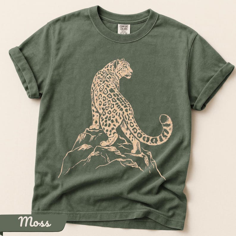 May include: A moss green short-sleeve t-shirt featuring a light tan graphic print of a leopard standing on a rocky outcrop. The shirt has a crew neckline and rolled sleeves, with a Comfort Colors brand label visible at the inside of the collar.
