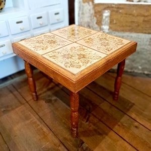 Könnte beinhalten: Kleiner, quadratischer Holztisch mit gefliester Oberfläche mit floralem Design. Der Tisch hat vier gedrechselte Beine und eine warme, braune Oberfläche. Die Fliesen sind hellbeige mit einem detaillierten, geprägten Muster.