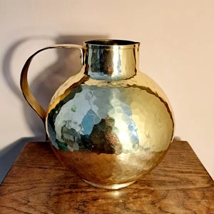 Può includere: Una brocca in ottone color oro lucido con corpo sferico e texture martellata. Il recipiente presenta una base ampia e arrotondata che si restringe verso un collo cilindrico liscio e include un singolo manico curvo fissato sul lato.