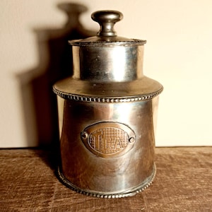 Peut inclure: Une théière argentée avec un couvercle et une petite poignée. La théière présente une plaque ovale avec le mot "TEA". La théière est posée sur une surface en bois. La théière mesure environ 15 cm de haut.