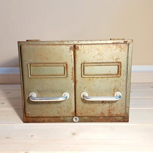 Op de afbeelding: Een vintage metalen archiefkast met twee laden, met een verweerde olijfgroene afwerking. Elke lade heeft een rechthoekige etikethouder en een gebogen chromen handgreep. De kast vertoont tekenen van roest en slijtage.