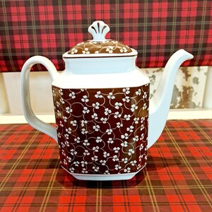 Könnte beinhalten: Eine weiße und braune Keramik-Teekanne mit floralem Muster. Die Teekanne hat einen gebogenen Ausguss, einen Henkel und einen Deckel mit dekorativem Knauf. Der Korpus der Teekanne ist braun mit weißem Blumendesign.