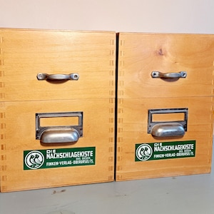 Könnte beinhalten: Zwei Holz-Aufbewahrungsboxen mit Metallgriffen und Etikettenhaltern. Jede Box hat einen grün-weißen Aufkleber mit deutschem Text. Die Boxen sind aus hellem Holz gefertigt und haben ein schlichtes, funktionales Design.