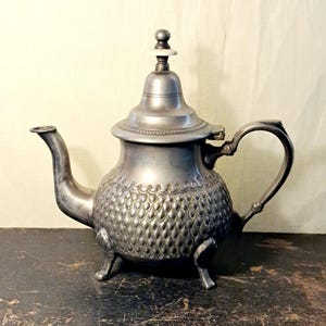 Könnte beinhalten: Eine metallische Teekanne mit mattgrauem Finish, die ein strukturiertes Schuppenmuster auf dem bauchigen Körper, einen geschwungenen Ausguss, einen Klappdeckel mit einem kleinen weißen Knauf und drei kurze, geschwungene Standfüße aufweist.