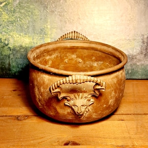 Könnte beinhalten: Ein verwitterter, bronzefarbener Metalltopf mit zwei verzierten Griffen mit einem Löwenkopf-Design. Der runde Topf hat eine weite Öffnung und steht auf einer Holzoberfläche. Der Hintergrund hat ein gemaltes, strukturiertes Aussehen.